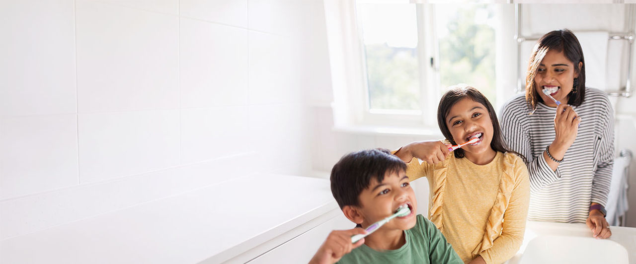 Indian mother and two children happily brushing their teeth together in a bright modern bathroom.