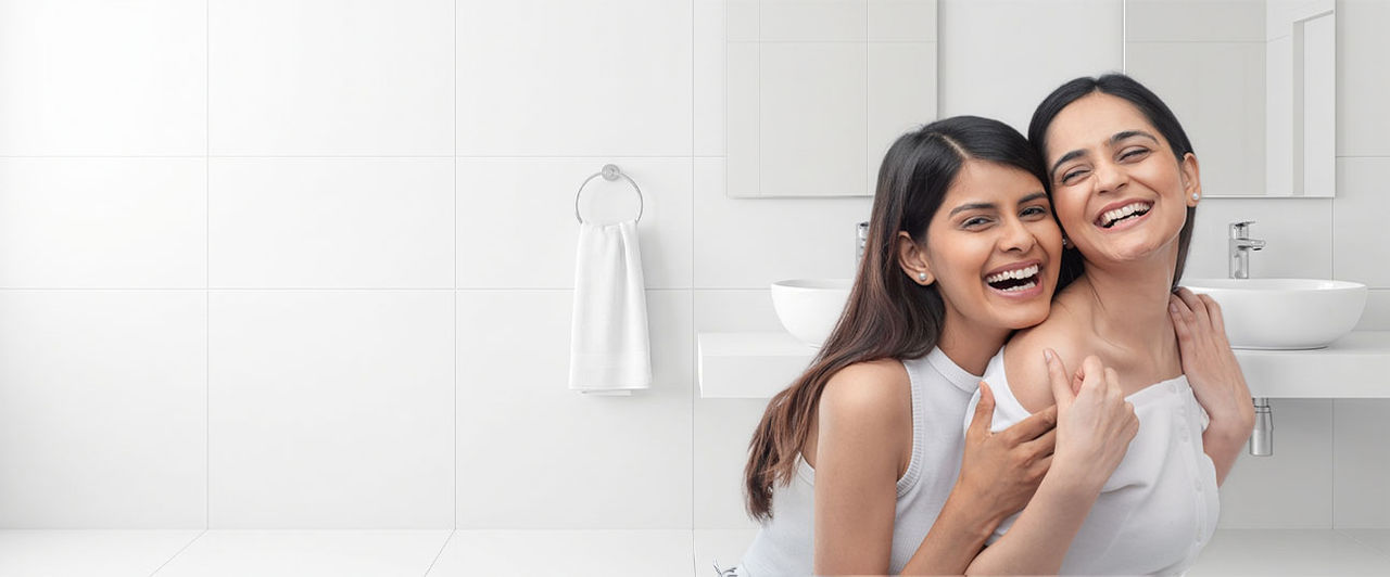 Two happy Indian women embracing and laughing together in a bright, modern white bathroom.
