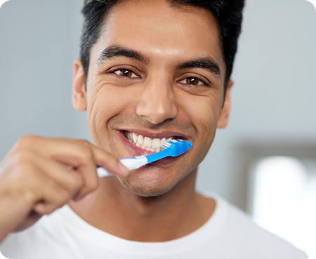 Close-up portrait of a smiling man with a white t-shirt brushing his teeth with a blue toothbrush in a home setting.