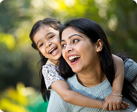 Joyful Indian mother laughing widely outdoors while giving her young daughter a piggyback ride in a sunlit park with a bokeh background.