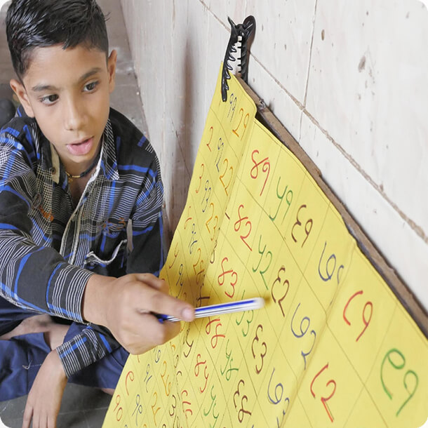 Young student pointing at a yellow number grid chart written in Devanagari numerals with a pen during a math lesson.